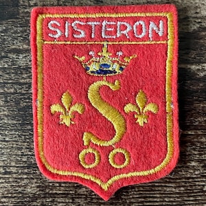 May include: A rectangular red embroidered patch with a gold border. The text SISTERON is stitched in white at the top. Below, a gold crown sits above a large gold letter S flanked by two gold fleur-de-lis symbols and two gold circles.