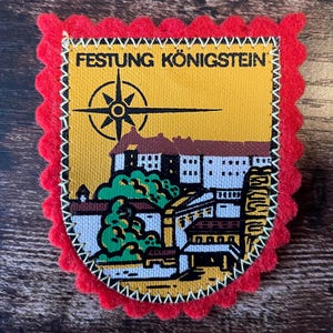 May include: A rectangular fabric patch with a scalloped red felt border. The centre features a yellow background with a black compass rose and a graphic illustration of Festung Königstein. The text FESTUNG KÖNIGSTEIN is printed in black at the top.