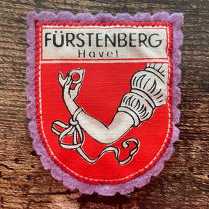 May include: A rectangular patch with a red background and a white and red emblem. The emblem features a white arm holding a ring, with the text "FÜRSTENBERG Havel" above. The patch has a scalloped purple border.