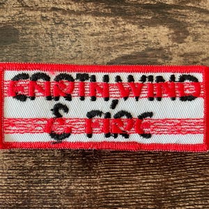 May include: Embroidered patch with the words "GORTH WIND & FIRE" in black and red lettering. The patch has a white background with red horizontal stripes and a red border. The patch is rectangular.