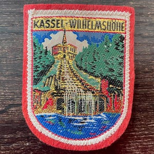 Könnte beinhalten: Ein rechteckiger gestickter Aufnäher mit rotem Rand und der Aufschrift KASSEL-WILHELMSHOHE am oberen Rand. Das Motiv zeigt ein mehrstöckiges Gebäude mit einem zentralen Turm, umgeben von grünen Bäumen und einem blauen Gewässer am Fuß.