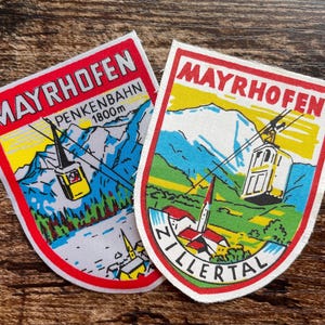 Könnte beinhalten: Zwei bestickte Stoffaufnäher mit dem Ortsnamen MAYRHOFEN in roter Schrift. Ein Aufnäher zeigt eine gelbe Seilbahn, Berge und den Text PENKENBAHN 1800m. Der andere zeigt eine Seilbahn, Berge und den Schriftzug ZILLERTAL am unteren Rand.