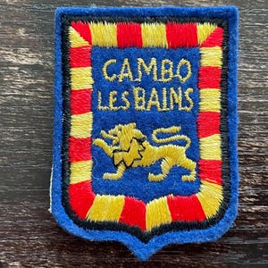 May include: A rectangular embroidered patch with a blue felt base. The center features a gold lion and the text CAMBO LES BAINS in gold thread. A border of alternating red and yellow vertical stripes surrounds the central blue field.