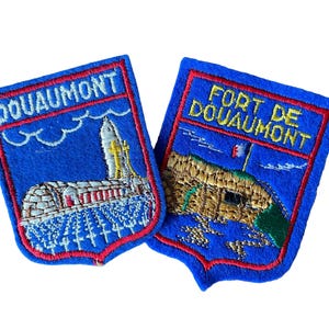 Puede incluir: Dos parches bordados azules con bordes rojos. Uno de los parches dice "DOUAUMONT" y representa un edificio y una torre. El otro parche dice "FORT DE DOUAUMONT" y muestra un fuerte con una bandera francesa. Ambos parches tienen forma de escudo.
