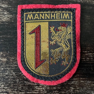May include: A red felt patch with a black and gold embroidered crest. The crest features a gold number "1" and a gold lion with a red crown on a black background. The text "MANNHEIM" is embroidered in gold above the crest.