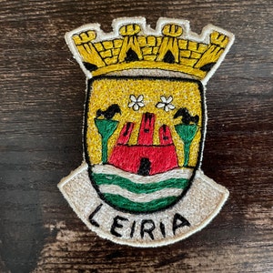Könnte beinhalten: Gestickter Aufnäher mit dem Wappen von Leiria, Portugal. Das Design umfasst eine rote Burg, grüne und weiße Wellen und den Text "LEIRIA". Der Aufnäher hat ein gelbes, weißes, grünes und rotes Farbschema.