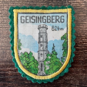 May include: A shield-shaped embroidered patch featuring a tall stone tower illustration, green trees, and a light blue sky. The text GEISINGBERG and 824m are printed at the top. The patch has a yellow inner border and a scalloped dark green edge.