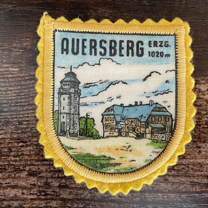 Puede incluir: Parche de Auerberg Erzgebirge con borde festoneado amarillo. El parche presenta una torre y un edificio con techo azul, sobre un fondo de cielo. El texto "AUERSBERG ERZG. 1020m" está impreso en la parte superior.