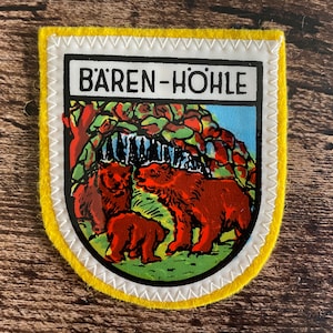 May include: A yellow-bordered patch with a white stitched edge. The patch features a graphic of three brown bears in front of a waterfall and trees. The text "BÄREN-HÖHLE" is displayed at the top in black letters.