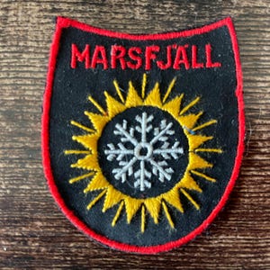 May include: A shield-shaped embroidered patch with a black background and a red border. The text MARSFJÄLL is stitched in red at the top. In the center, a yellow sunburst design surrounds a light gray snowflake icon with intricate details.