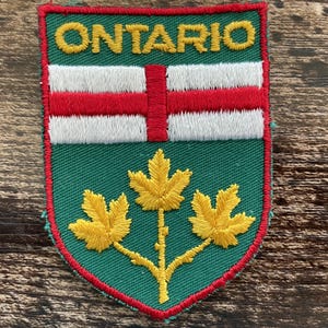 Op de afbeelding: Een schildvormige geborduurde patch met het woord ONTARIO in gele draad aan de bovenzijde. Het ontwerp bevat een horizontale witte band met een centraal rood kruis op een groene achtergrond en drie gele esdoornbladeren onderaan.