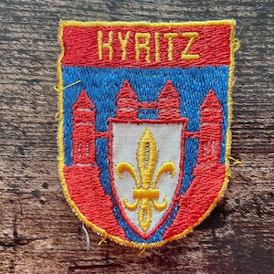Peut inclure: Un écusson brodé en forme de blason avec le texte KYRITZ en fil jaune sur fond rouge. Le motif présente un champ bleu avec des tours rouges et un écu central blanc contenant une fleur de lys jaune, le tout bordé de fil jaune.