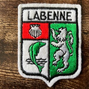Peut inclure: Un écusson brodé en forme de blason portant l'inscription LABENNE en haut. Le motif comprend une section rouge avec une coquille Saint-Jacques blanche, une section blanche avec un poisson vert au-dessus de vagues, et une section verte avec un lion argenté.