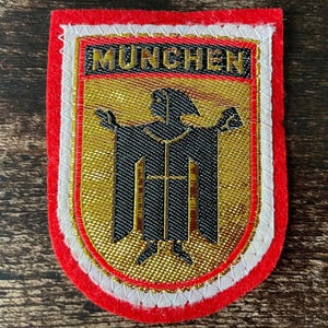 Könnte beinhalten: Ein rechteckiger gestickter Aufnäher mit rotem Filzrand und weißem Stickrand. In der Mitte befindet sich ein goldener Schild mit einer schwarzen Mönchsfigur in einer Robe, darüber das Wort MUNCHEN in schwarzen Buchstaben auf Gold.