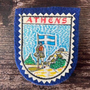 Puede incluir: Un parche de fieltro azul con un borde blanco festoneado que presenta una ilustración gráfica de una persona con vestimenta tradicional sosteniendo una bandera griega. El diseño incluye la Acrópolis y la palabra ATHENS en letras rojas.