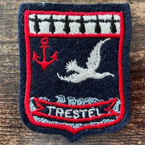 Op de afbeelding: Een marineblauw, schildvormig geborduurd embleem met een rood anker, een witte vogel in vlucht en een witte band met zwarte hermelijnvlekken aan de bovenzijde. Een rode banner onderaan bevat de witte tekst TRESTEL, omlijnd met rood borduurwerk.