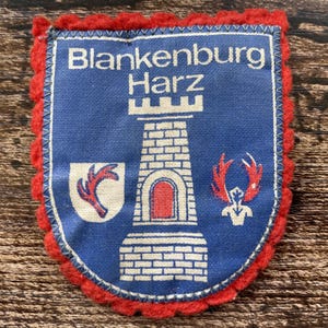 Può includere: Una toppa in tessuto blu con bordo smerlato rosso che presenta il testo bianco Blankenburg Harz, un'illustrazione centrale di una torre in pietra bianca, uno scudo bianco con un ramo rosso e un emblema di corna di cervo rosso e bianco.