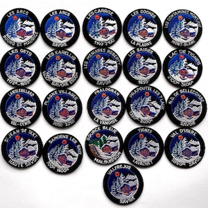 May include: A collection of twenty-one circular embroidered patches featuring a winter mountain scene with a chalet, pine trees, and a sun. Each patch displays a unique French ski resort name and elevation text in white thread on a black border.