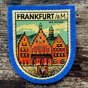Könnte beinhalten: Ein blauer Filzaufnäher mit einer grafischen Darstellung des Frankfurter Römer-Gebäudekomplexes. Der Text FRANKFURT/a.M. und DER ROMER ist oben aufgedruckt. Das Design umfasst orangefarbene Gebäude, ein grünes Dach und ein kleines schwarzes Wappen.