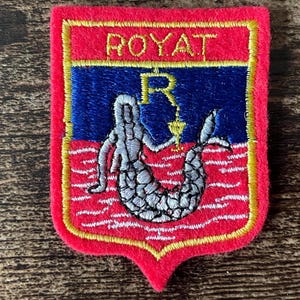 May include: A shield-shaped embroidered patch with a red border and background. The top section features the text ROYAT in yellow. Below, a blue field contains a yellow letter R above a silver mermaid figure, set against a red base with white waves.
