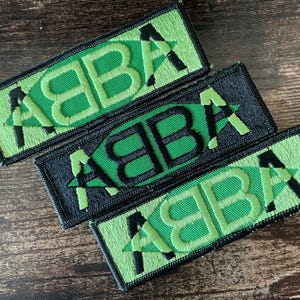 以下が含まれることがあります： 緑と黒で「ABBA」の文字が刺繍された3つのパッチ。パッチは長方形で、文字は様式化されたフォントです。2つのパッチは緑色の背景で、1つは黒色です。