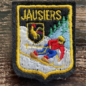 May include: A rectangular embroidered patch with a black background and a yellow border. It features the text JAUSIERS in yellow, a shield with a rooster, a skier in a red jacket and blue pants on a white snowy slope, and green pine trees.