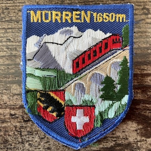 Op de afbeelding: Een schildvormige geborduurde patch met de tekst MURREN 1650m in het geel. Het ontwerp toont een rode trein op een stenen viaduct, besneeuwde bergen, groene dennenbomen en twee heraldische schilden, waarvan één met een wit kruis op rood.