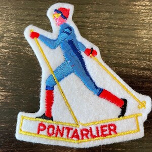 Peut inclure: Un écusson brodé sur fond de feutrine blanche représentant un skieur de fond vêtu d'une combinaison bleue, de chaussettes rouges et d'un bonnet rouge. Le skieur tient des bâtons jaunes au-dessus d'une bannière à bordure jaune avec le texte PONTARLIER.