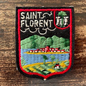 以下が含まれることがあります： 黒い背景と赤い縁取りの盾型刺繍ワッペン。白い文字でSAINT FLORENTと記され、緑の木と白い塔が描かれた小さな紋章、緑の丘、青い水面、黄色い畑が刺繍された風景画がデザインされています。