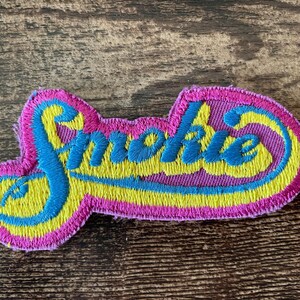 Może przedstawiać: Haftowana naszywka ze słowem "Smokie" w stylu retro. Naszywka ma różową obwódkę, żółte i niebieskie akcenty oraz niebieską czcionkę. Naszywka jest prawdopodobnie przeznaczona na odzież lub akcesoria.