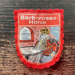 Puede incluir: Parche rojo y blanco con el texto "Barbarossa-Höhle" y una ilustración de una figura coronada con una máscara roja. El parche tiene un borde rojo festoneado, probablemente un recuerdo.