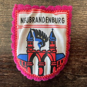 May include: A fabric patch with the word NEUBRANDENBURG at the top. The patch features a red and blue illustration of a building with arches and towers. The patch has a pink, scalloped border.