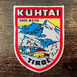 Könnte beinhalten: Ein rechteckiger gestickter Aufnäher mit abgerundeten unteren Ecken, der eine Berglandschaft zeigt. Der Text KUHTAI steht oben, 2000 METER darunter und TIROL am unteren Rand. Das Motiv zeigt ein gelbes Gebäude mit blauem Dach vor blau-weißen Bergen, alles umrandet von einem dicken roten Rahmen.