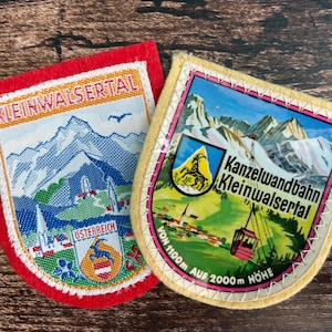 Könnte beinhalten: Zwei gestickte Souvenir-Aufnäher. Der linke Aufnäher hat einen roten Rand, den Schriftzug KLEINWALSERTAL, eine Berglandschaft und ein OSTERREICH-Wappen. Der rechte Aufnäher hat einen gelben Rand, den Text Kanzelwandbahn Kleinwalsertal VON 1100m AUF 2000m HÖHE, eine Bergkulisse, eine Seilbahn und ein Wappen mit einem Steinbock.