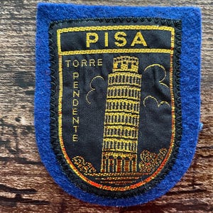 Könnte beinhalten: Ein blauer Filzaufnäher mit einer schwarzen, bestickten Schildform und goldenen Nähten. Der Text PISA und TORRE PENDENTE ist in Gold gestickt, zusammen mit einer detaillierten goldenen Darstellung des Schiefen Turms von Pisa mit Wolken.