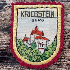Puede incluir: Un parche de tela rectangular con un borde de fieltro rojo oscuro festoneado y un borde bordado en amarillo. El centro presenta una ilustración impresa del castillo de Kriebstein con techos rojos, paredes blancas y árboles verdes, con el texto KRIEBSTEIN BURG.