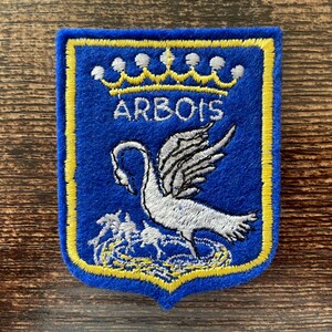 May include: A blue embroidered patch in a shield shape with a yellow border. It features a silver swan above a yellow nest, topped by a yellow crown and the text ARBOIS in white embroidery. The design is centred on a blue felt-like fabric base.