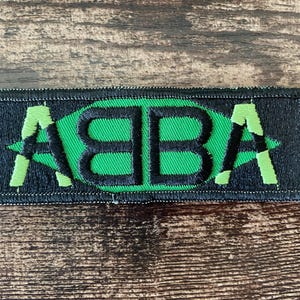 Puede incluir: Un parche rectangular con la palabra "ABBA" bordada en verde sobre un fondo negro. Las letras están estilizadas y sobre una forma ovalada verde. El parche tiene un borde negro y es probablemente para planchar o coser.