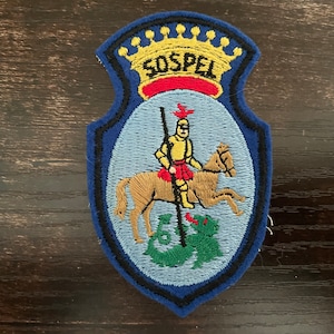 May include: A shield-shaped embroidered patch featuring a blue border and a light blue centre. At the top, a yellow crown sits above the text SOSPEL in black. Below, a knight in yellow armour rides a brown horse, piercing a green dragon with a spear.