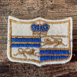 May include: A shield-shaped embroidered patch featuring a white felt base with gold metallic thread borders. The design includes a blue crown at the top, with alternating quadrants of blue horizontal stripes and a gold lion figure on a white background.