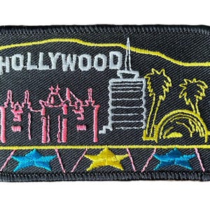 Könnte beinhalten: Ein rechteckiger, schwarz bestickter Aufnäher mit dem weißen Schriftzug HOLLYWOOD über einer rosa Skyline, einem weißen Turm, gelben Palmen und einem gelben Bogen. Der untere Rand enthält zwei blaue und einen gelben Stern auf schwarzem Grund.