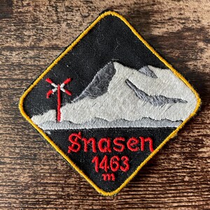 May include: A diamond-shaped embroidered patch with a black background and a yellow border. It features a white and grey mountain peak, a red cross-shaped symbol, and the red embroidered text Snasen 1463 m.