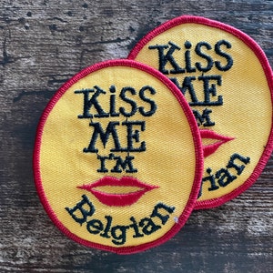 Puede incluir: Dos parches redondos amarillos con ribete rojo. Cada parche presenta el texto "Kiss Me I'm Belgian" en negro, con un gráfico de labios rojos. Los parches son probablemente para ropa o accesorios.