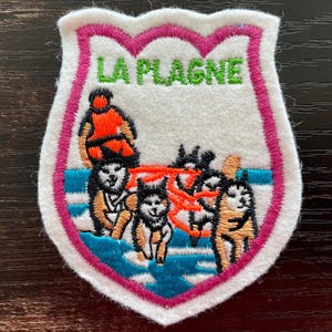 Op de afbeelding: Een schildvormige geborduurde patch met een witte achtergrond en een roze rand. Het bevat de groene tekst LA PLAGNE boven een illustratie van een persoon in een oranje jas die een hondenslee bestuurt door een blauw besneeuwd landschap.