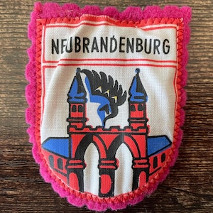 May include: A shield-shaped fabric patch with a bright pink scalloped border. The center features a white background with the text NEUBRANDENBURG in black. Below the text is a graphic of a red gatehouse with blue roofs and a black bird.