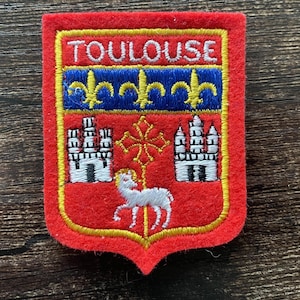 以下が含まれることがあります： 「TOULOUSE」の文字が金色のユリの紋章が付いた青いバナーの上に刺繍されたパッチ。パッチは赤い背景に金色の縁取りがあり、白い子羊と城のデザインが含まれています。
