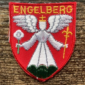 Könnte beinhalten: Ein schildförmiger gestickter Aufnäher mit rotem Hintergrund und gelbem Rand. Der Text ENGELBERG ist oben in Gelb gestickt. Ein weißer Engel mit Flügeln hält eine weiße Blume und einen gelben Stab und steht auf einem grünen Sockel.