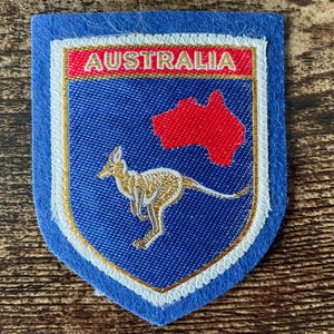 Peut inclure: Un écusson brodé en forme de bouclier avec une bordure en feutre bleu. Le centre présente un fond bleu avec une silhouette de kangourou dorée, une carte rouge de l'Australie et une bannière rouge avec le texte blanc AUSTRALIA en bordure dorée.
