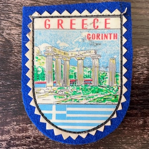 May include: A blue felt souvenir patch featuring a white scalloped border. The center graphic depicts the ancient ruins of Corinth, Greece, with the text GREECE and CORINTH printed above. The bottom section displays the blue and white Greek flag.