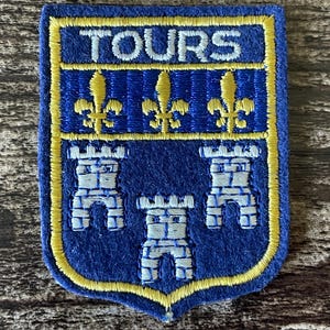 May include: A shield-shaped embroidered patch with a blue background and yellow border. The word TOURS is stitched in white at the top. Below, three yellow fleur-de-lis symbols are arranged horizontally, followed by three white stone towers.